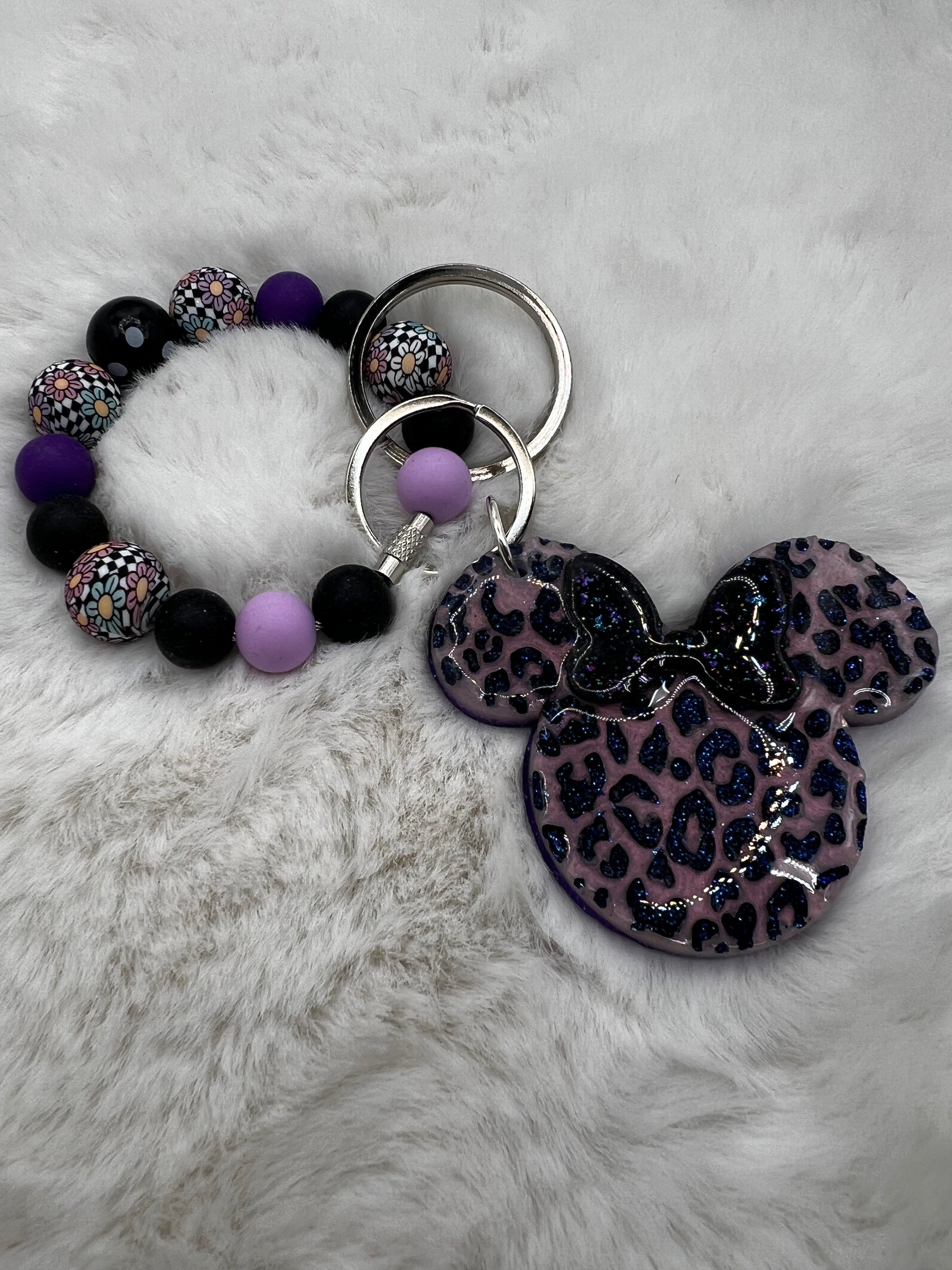 Keychain with leopard print Minnie Mouse pendant and a beaded ring with purple and black beads on a fluffy white surface.