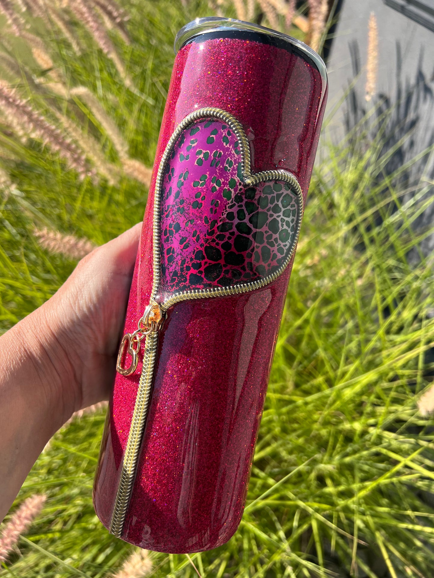 Red glittery tumbler with a gold zipper held in front of green grass