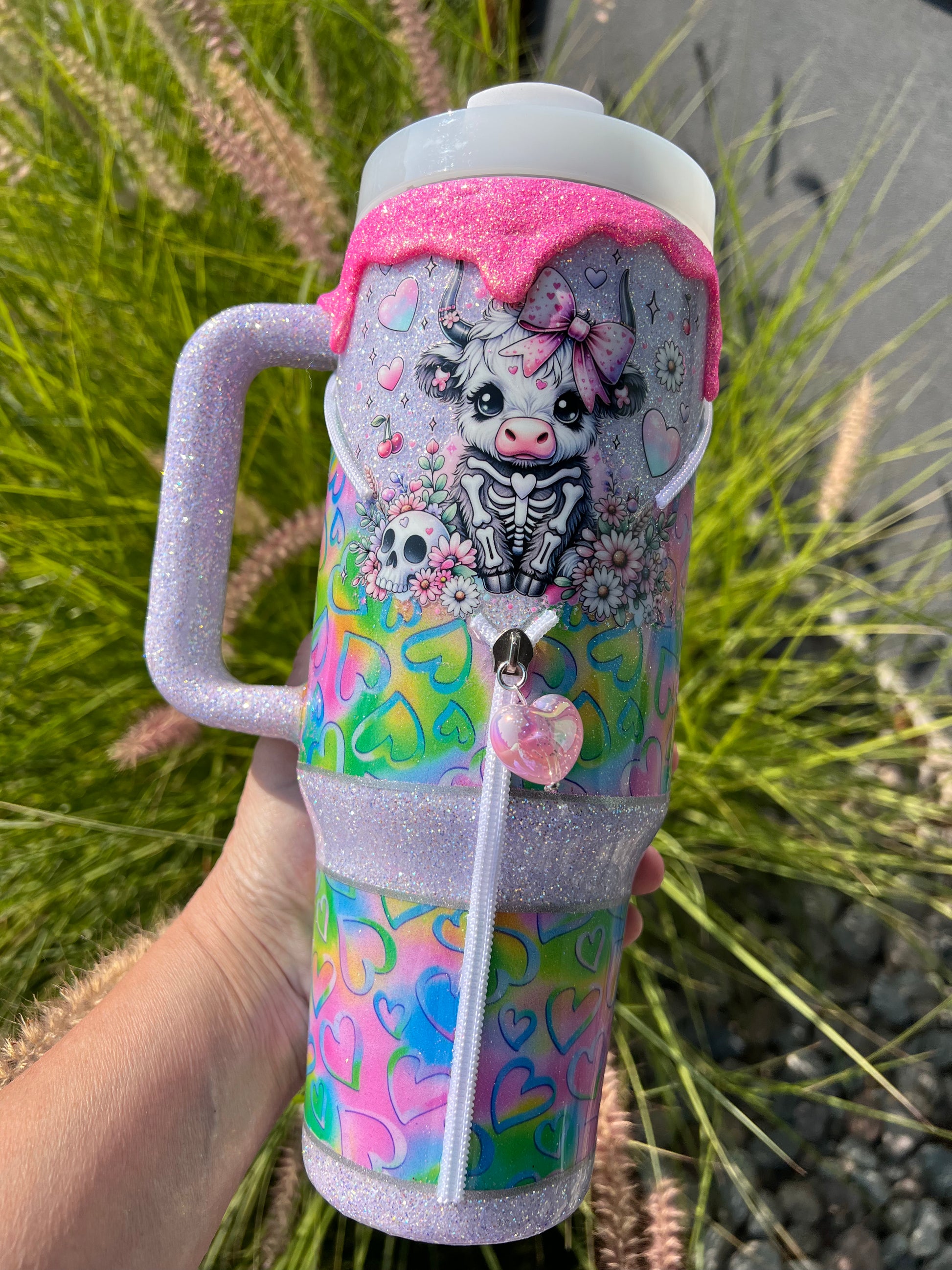 Colorful insulated tumbler with a design of a cow and hearts held by a hand against a natural background.