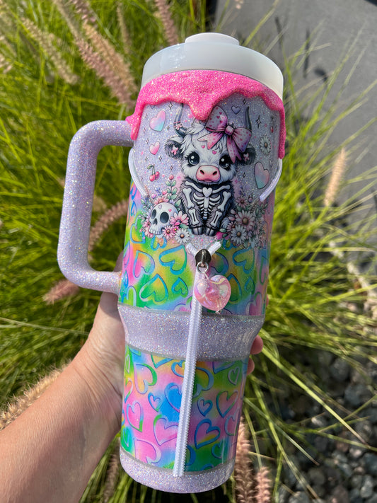 Colorful insulated tumbler with a design of a cow and hearts held by a hand against a natural background.