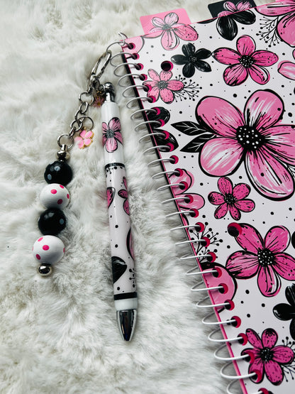 Pen with decorative keychain and floral notebook on a white surface
