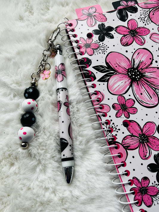 Pen with decorative keychain and floral notebook on a white surface