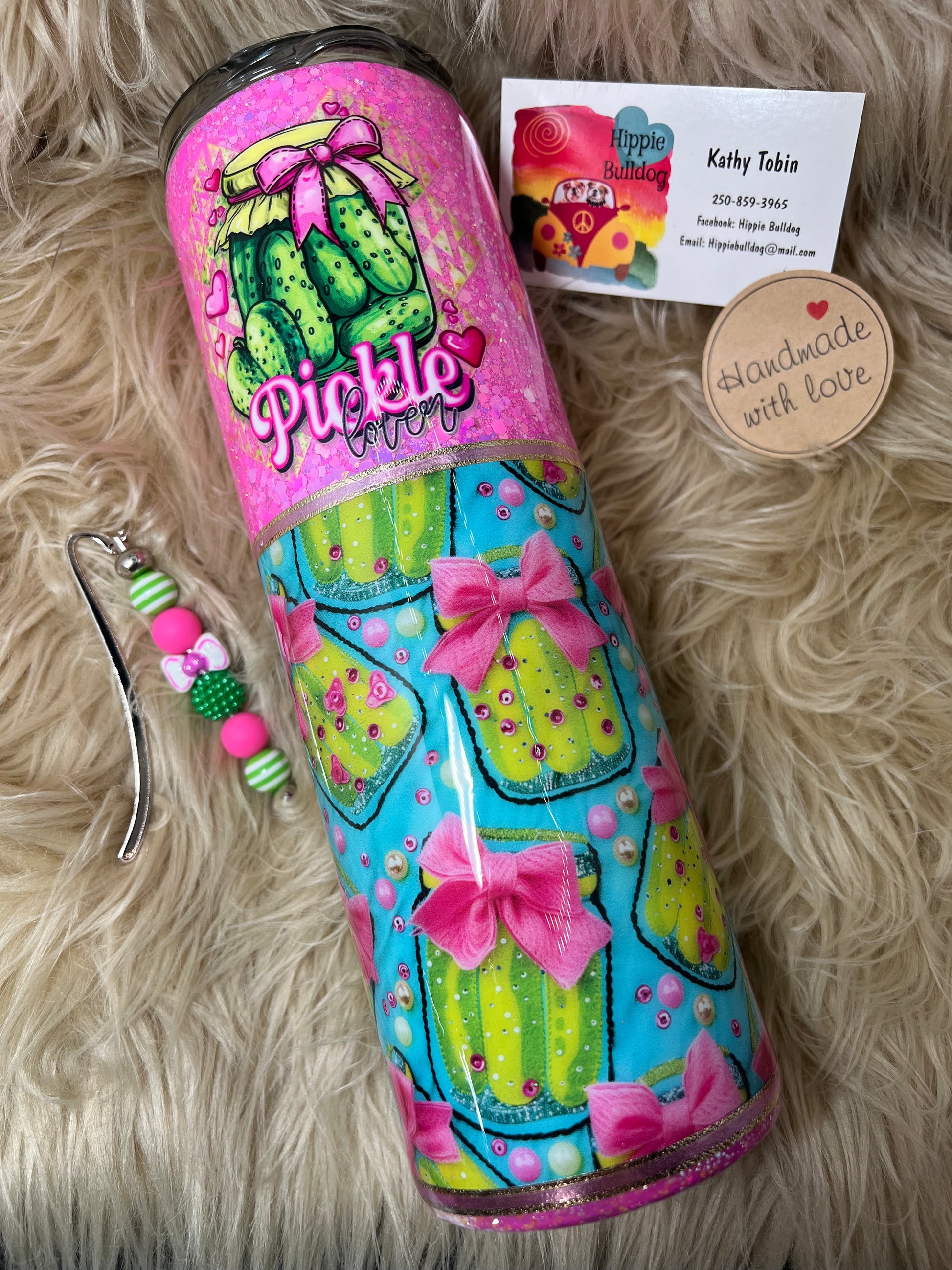 Colorful tumbler with pickle design on a fluffy surface, accompanied by a charm and business card.