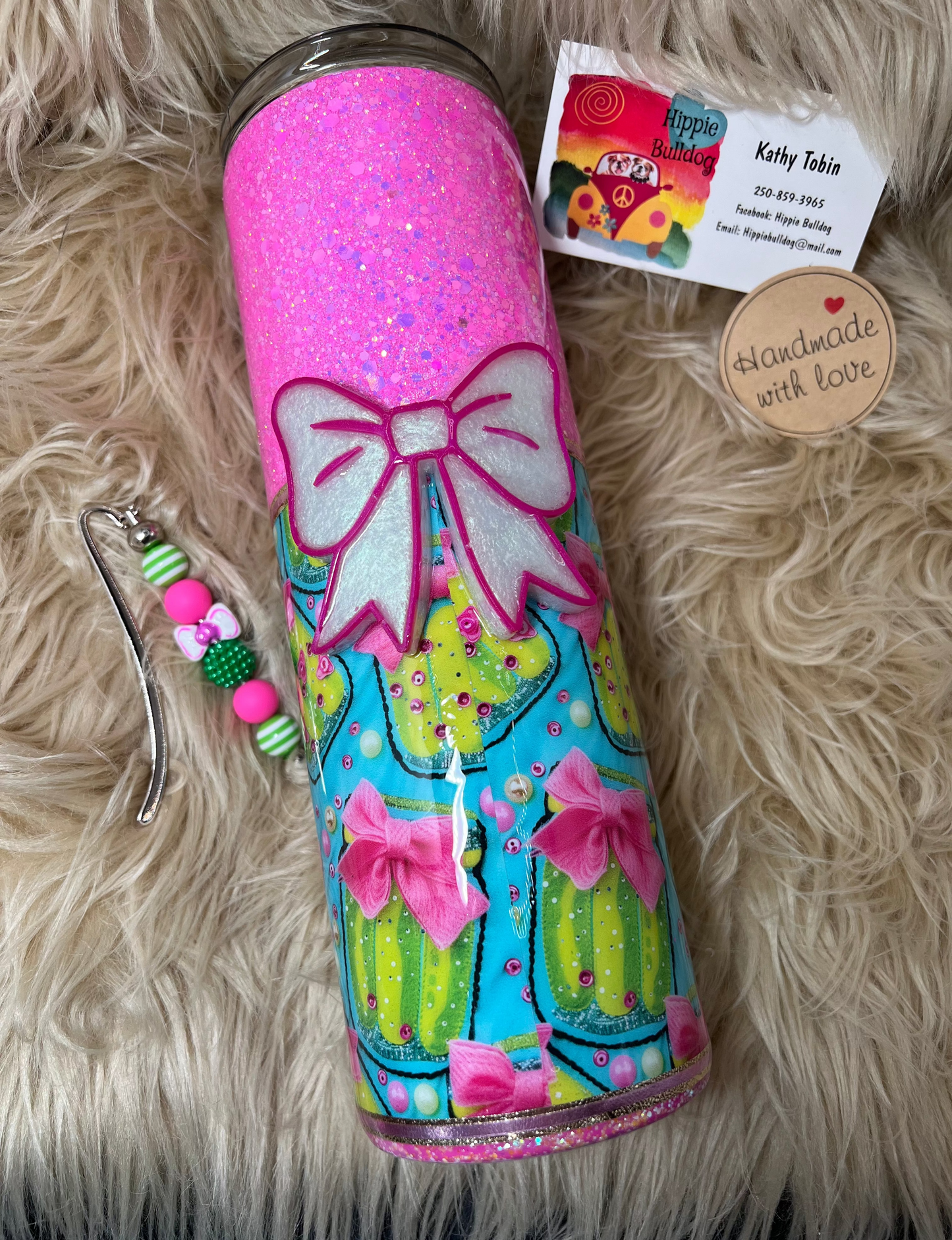 Colorful tumbler with a pickle theme and bow design on a fluffy surface, accompanied by a business card and decorative charms.
